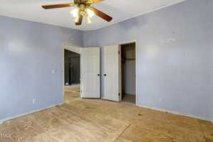 An empty room with light purple walls and a plywood floor. There is a ceiling fan with lights, a white door leading to another room, and a closet with a door slightly open. Some walls show signs of wear.