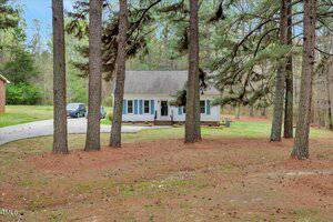 A small white house with blue shutters is surrounded by tall pine trees. A driveway leads to the house, where a black vehicle is parked. The lawn is grassy with patches of pine needles, and the area is wooded and quiet.