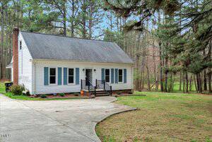A white two-story house with a dark shingle roof and blue shutters, surrounded by trees. It features a small front porch with black railings and a chimney on the left. The house is situated along a curved driveway amidst a grassy lawn.