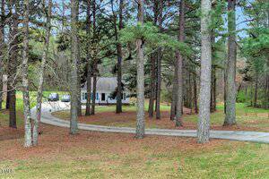 A house with a blue exterior is partially visible through tall pine trees. A curved driveway leads to the house, with a few parked cars. The surrounding area is grassy with patches of pine straw. The sky is overcast.