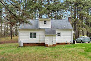A small white house with a gray shingled roof is set among tall pine trees. It has a single dormer window and a central back door. A silver car is parked on the right side. The grass lawn is expansive and green.