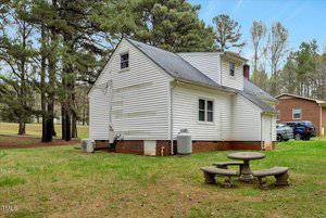 A white two-story house with a brick foundation sits in a grassy yard. Tall pine trees are visible in the background. There is a stone picnic table with benches on the lawn. Two vehicles are parked beside the neighboring house in the distance.