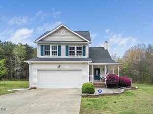 Nestled on 3690 Mabel Lane, this charming two-story house boasts a gray exterior with blue shutters. It features an attached garage and a cozy front porch. The yard is adorned with a curved walkway, manicured bushes, and an American flag waving under the clear sky with trees as a backdrop.