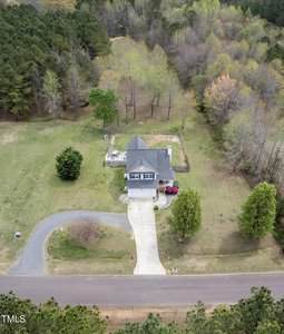 Aerial view of a two-story house with a gray roof, situated in a large grassy area. A driveway leads to the house, which has a fenced backyard. Surrounding the property are numerous trees, with a road running in front of the house.