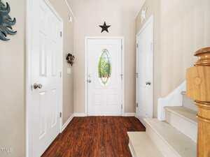 A hallway with a wooden floor features a white front door with an oval glass panel. The walls are light-colored, with two additional white doors on either side. A black star decoration is above the front door. A wooden staircase is on the right.