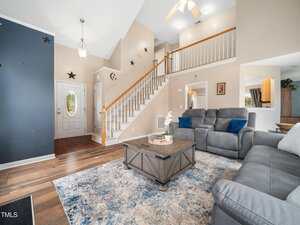 A spacious living room featuring a staircase with white railings and wood steps. It has a gray couch, a patterned rug, and a wooden coffee table with decorative items. A ceiling fan and chandelier hang above. There are star decorations on the walls.
