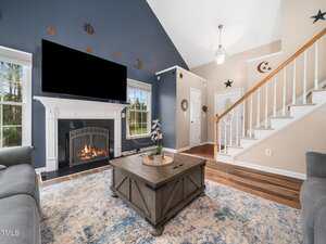 A living room with a vaulted ceiling features a fireplace under a wall-mounted TV. There's a wooden coffee table with a plant, surrounded by gray sofas. The walls have star and moon decorations, and a staircase leads upstairs by the entrance door.