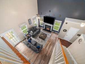 A spacious living room with a high ceiling, viewed from above. It features two gray couches, a coffee table, and a fireplace with a TV above it. The room has wooden flooring, a large rug, and several windows, allowing natural light to enter.