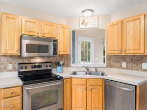 Kitchen with light wood cabinets and beige countertops. Features a stainless steel stove, microwave, and dishwasher. A double sink is below a small window. Beige tile backsplash. A small decorative sign above the sink reads "Home Sweet Home.