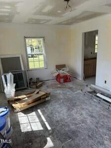 Unfinished room with unpainted walls and a bare floor. Includes a window, various construction materials, tools, a ladder, paint cans, and a partially assembled frame leaning against the wall. Natural light enters through the window.