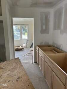 View of a partially completed room under renovation with visible drywall patches. Wooden cabinets are installed on the right, with unfinished countertop space above. Construction materials and tools are visible in adjacent room through the doorway.