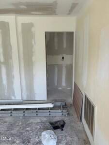 Room under construction with drywall partially installed. Walls have visible joint compound and patches. The floor is bare, with construction materials scattered. A small door opening leads to another space. An electrical outlet is midway on the wall.