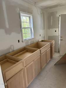 Unfinished kitchen with light wood base cabinets installed, but no countertop or appliances. Walls are partially primed, and a window allows natural light. An open door and an electrical socket are visible.