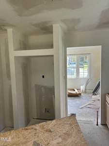 A room under construction with partially finished drywall. An alcove with utility hookups is visible, possibly for laundry. A door leads to another room with construction materials on the floor and sunlight coming through windows.