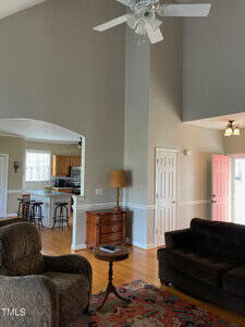 Living room interior with high ceiling, beige walls, and hardwood floors. Includes a ceiling fan, dark sofa, patterned rug, and armchair. Visible kitchen with bar stools and a partial view of an open pink front door.