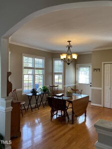 A dining area with wooden flooring features a drop-leaf table and several chairs. Large windows allow natural light to illuminate the space. A chandelier hangs overhead, and decorative items, including a wall sign reading "FAITH," are present.