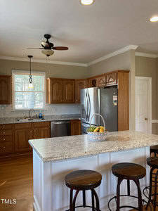 A kitchen with wooden cabinets, a stainless steel refrigerator, and a granite island with three bar stools. A basket of fruit sits on the island. A window above the sink lets in natural light. A ceiling fan is visible above.