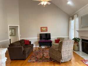 A living room with a high ceiling features a couch, two patterned chairs, a TV on a stand, and a colorful rug. There are potted plants, a fireplace, and a ceiling fan. The walls are light gray and a window with white curtains is on the right.