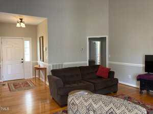Living room with a brown sofa, red and white cushions, and a patterned ottoman. A small TV is on a stand to the right. There is a console table near the white front door. Hardwood floors and a light fixture on the ceiling are visible.
