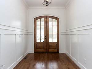A hallway with a wooden floor leads to a set of double wooden doors with glass panels. The walls are white with wainscoting, and there is a black metal light fixture hanging from the ceiling.