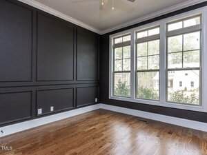 A room with dark paneled walls and large windows overlooking a house and trees. The room has a wooden floor, and there are electrical outlets on one wall. The windows let in natural light.