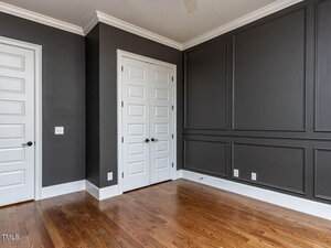 An empty room with dark gray walls, white double doors, and a light hardwood floor. The room has white trim and wall paneling. There are electrical outlets near the baseboards.