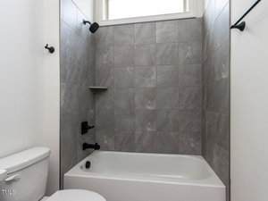 A bathroom with a white bathtub, gray tile wall surround, and a black showerhead. The toilet is partially visible on the left. A narrow window is above the shower, and white walls and a black towel rod are on the right.