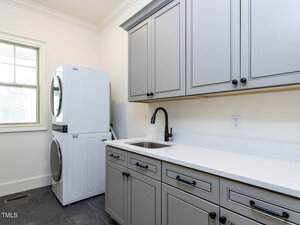 A laundry room with gray cabinets and a white countertop. There is a stainless steel sink with a black faucet. A stacked white washer and dryer are situated next to a window on the left side of the room.