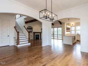 A spacious open-concept living area with hardwood floors, white walls, a staircase with wooden railings, a modern chandelier, a fireplace, and a kitchen with large windows and pendant lighting.