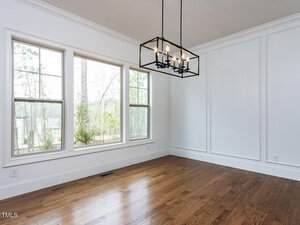 Bright, empty room with hardwood floors, three large windows, white walls with decorative molding, and a modern rectangular black chandelier hanging from the ceiling.