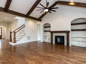A spacious living room with wood flooring, exposed dark wooden ceiling beams, a ceiling fan, built-in shelves, a fireplace with a stone surround, and a staircase leading upstairs. Walls are painted white.