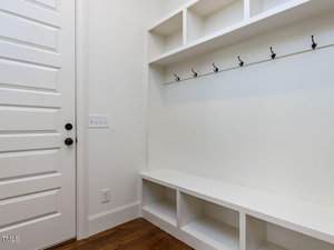 A white mudroom features open cubby shelves above and below, a row of black hooks for hanging items, and a bench with more storage. The room has wooden flooring, a white paneled door with two black knobs, and a light switch on the wall.