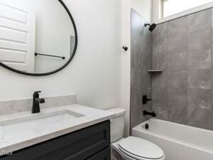 A modern bathroom features a white sink with a black faucet set in a dark vanity, a round mirror above, and a toilet beside it. A bathtub with gray tiled walls and black fixtures is on the right, with a small window above.