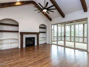 A spacious living room features a stone fireplace with built-in shelves on both sides, wooden ceiling beams, a modern ceiling fan, hardwood floors, and large sliding glass doors leading to a screened porch.