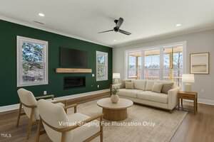 A living room with a green accent wall features two windows, a wall-mounted TV, and a fireplace. Furnished with a beige sofa, wooden chairs, and a coffee table. Ceiling fan and recessed lighting present. Large windows with views of trees.
