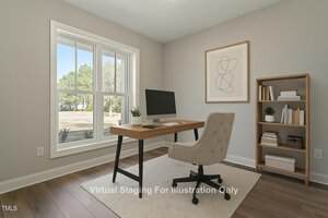 A home office features a wooden desk with a computer monitor and a beige swivel chair. A large window allows natural light in. A bookshelf with decor is on the right. The walls are light gray with a minimalist framed artwork hung above the desk.