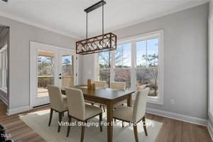 A dining room with six cream-colored chairs around a wooden table. A geometric chandelier hangs above. Glass doors lead to a wooden deck outside. Large windows offer views of trees. Text states "Virtual Staging For Illustration Only.