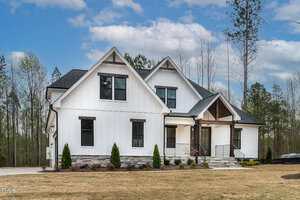 White modern farmhouse with black trim, featuring a gable roof and stone accents. The front has several windows, a porch with stairs, and landscaped shrubs, surrounded by a lawn. Trees are visible in the background under a blue sky with clouds.