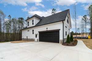 A modern two-story house with white siding and black roof and garage door. It has a paved driveway and a small landscaped area with shrubs. Trees are visible in the background under a partly cloudy sky.