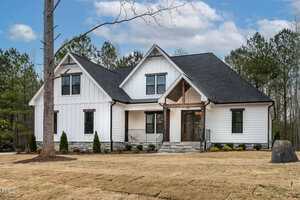Nestled at 3912 Cedar Knolls Drive in Youngsville, this white two-story house features a black roof, large windows, and a charming covered front porch. Stone steps lead to the entrance, surrounded by a neatly trimmed lawn and several small shrubs, with trees and a cloudy sky in the background.