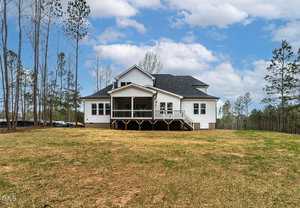 A two-story white house with a dark roof is situated in an open area surrounded by tall, sparse trees. The house features a back porch with stairs leading to a grassy backyard. The sky is partially cloudy.