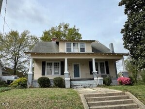 Located at 121 W Young Avenue in Henderson, this charming two-story house boasts pale yellow siding and a gray roof. A covered front porch with white columns, dark shutters, and a central chimney complements the inviting facade. The lawn features small bushes and a blooming pink bush on the right.
