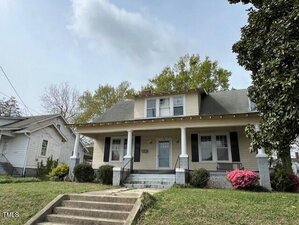 A two-story house with a covered front porch, featuring four columns and a set of stairs leading up to the entrance. The exterior is light-colored with dark shutters. Surrounding the house are trees, shrubs, and pink flowers. A sidewalk runs in front.