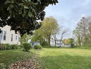 A suburban backyard with grass and several trees. A white house with siding is partially visible on the left, and there is a small blue shed in the middle distance. More houses and trees are seen in the background under a cloudy sky.