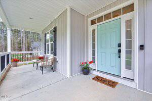 A front porch with a light blue door flanked by glass panels. Two chairs and a small table with red flowers are on the left. A doormat reads "Home Sweet Home." The porch overlooks trees and a neighboring house.