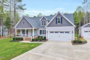 Single-story house with a gray gabled roof and white siding, featuring a porch with columns and a two-car garage. The driveway is concrete, and the yard is landscaped with shrubs. Surrounded by trees, the setting is suburban and wooded.