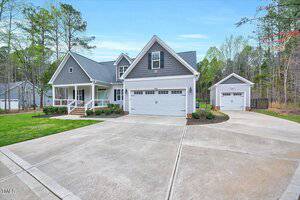A two-story gray house with white trim features a large driveway and a three-car garage. The property is surrounded by trees, and a basketball hoop is visible on the right. The front porch is accented with white railings and small shrubs.