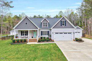 Located at 1165 Shonele Lane, this modern, single-story home boasts a gray exterior with white trim and a gable roof. It features a welcoming front porch, manicured lawn, and two-car garage. Surrounded by trees, the property also includes a paved driveway for easy access.