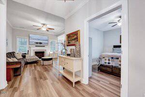 A hallway with light wood flooring leads to a living room on the left and a bedroom on the right. The living room has a sofa, fireplace, and wall-mounted TV. The bedroom has a bed with checkered bedding and a ceiling fan. A console table is in the hallway.