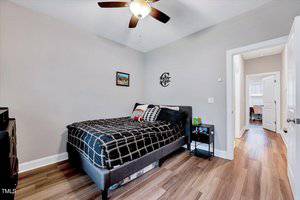 A bedroom with a gray bed frame and black checkered bedding. A ceiling fan with lights is above. A small black side table holds a clock. A framed picture hangs on the wall, and an open door leads to another room with wood flooring.